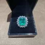 Bague en or blanc 18 carats sertie d’une émeraude d’environ 5 carats entourée de diamants, présentée dans son écrin.
